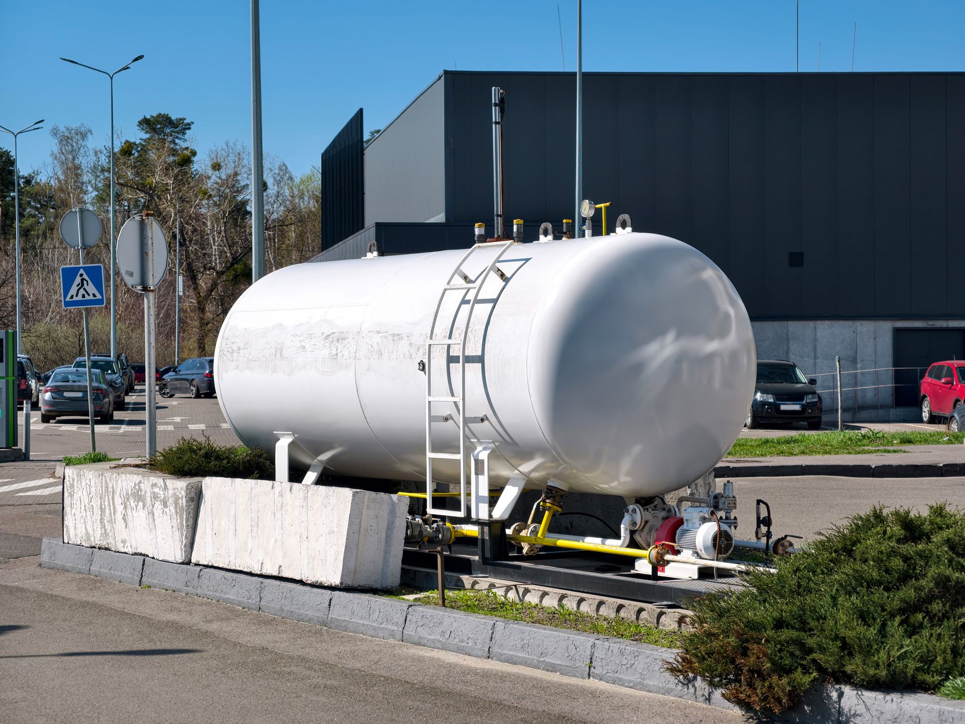 White cylindrical tank on a trailer beside a road and industrial building, with a ladder attached
