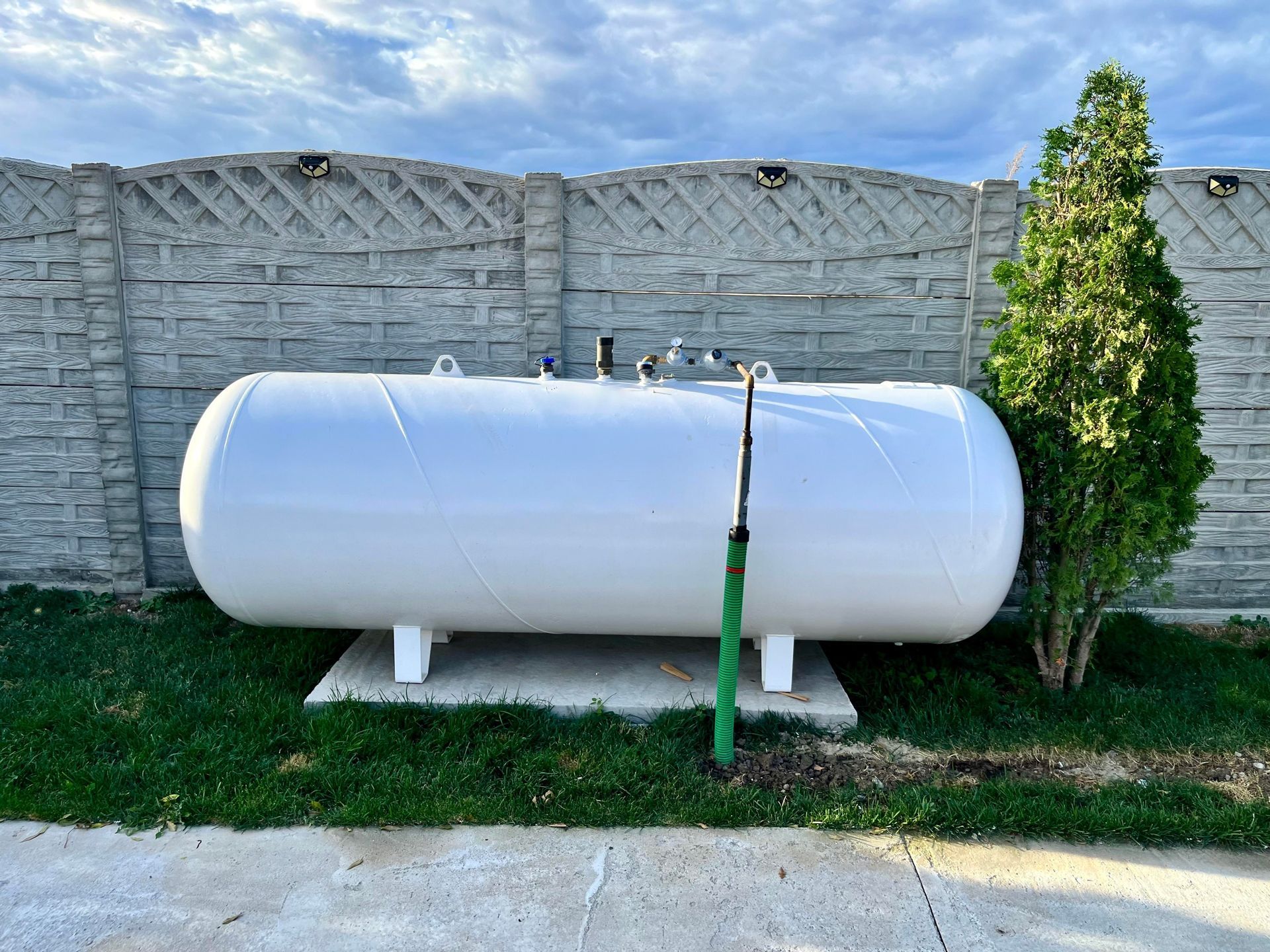 White propane tank on a grassy yard beside a stone fence and green shrub.