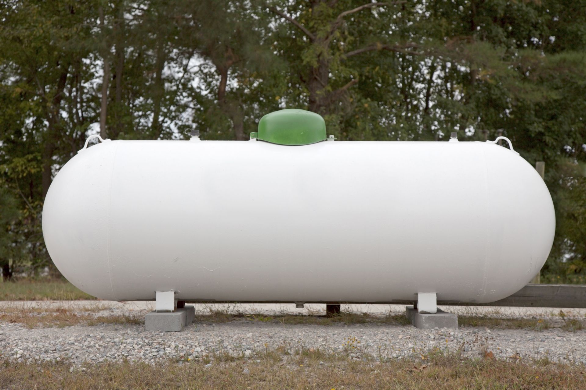 White horizontal propane tank with a green dome valve on gravel, with trees in the background