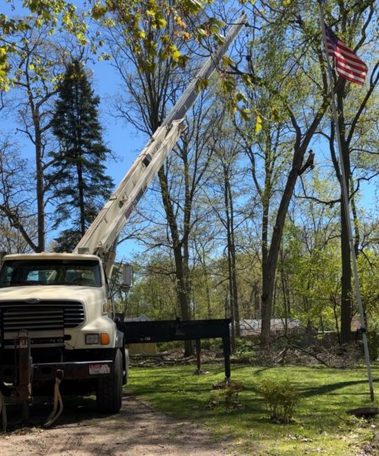 Truck-mounted crane hoisting a tall pole beside trees, with an American flag in the yard