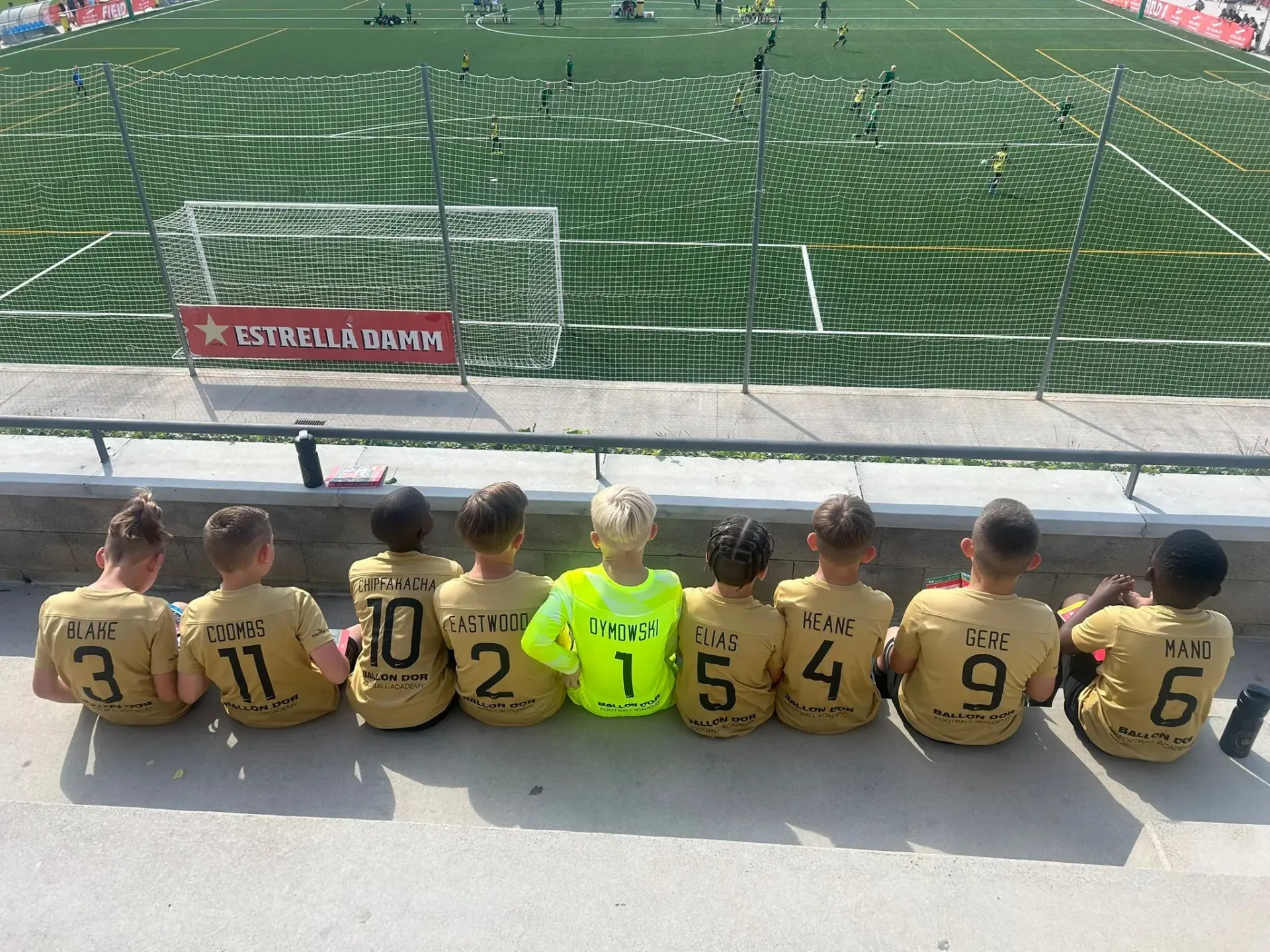 Children in soccer jerseys sit on a bench, facing a fenced soccer field with green turf.