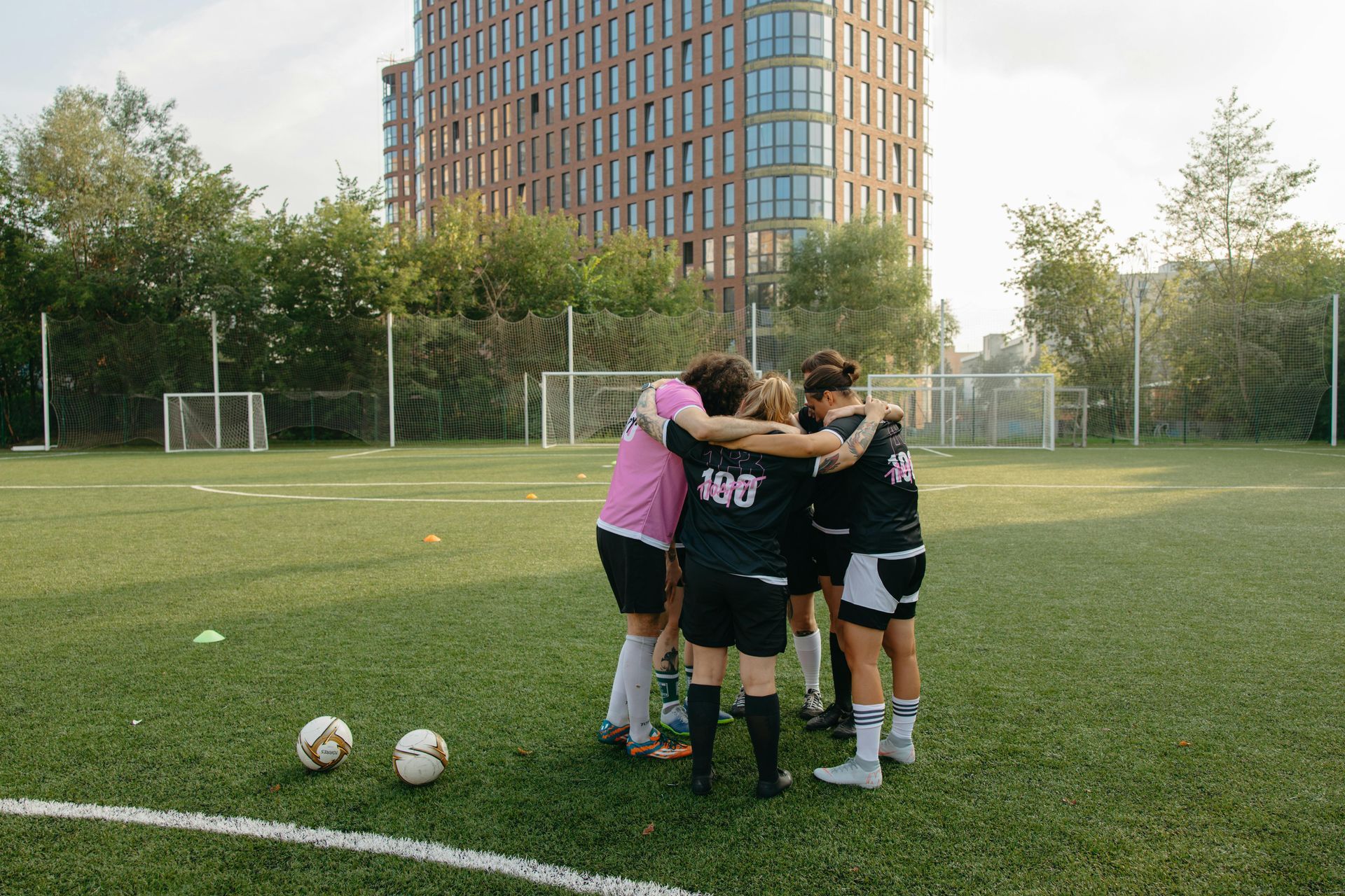 Soccer players huddle on a field near two balls, with a tall building and trees in the background