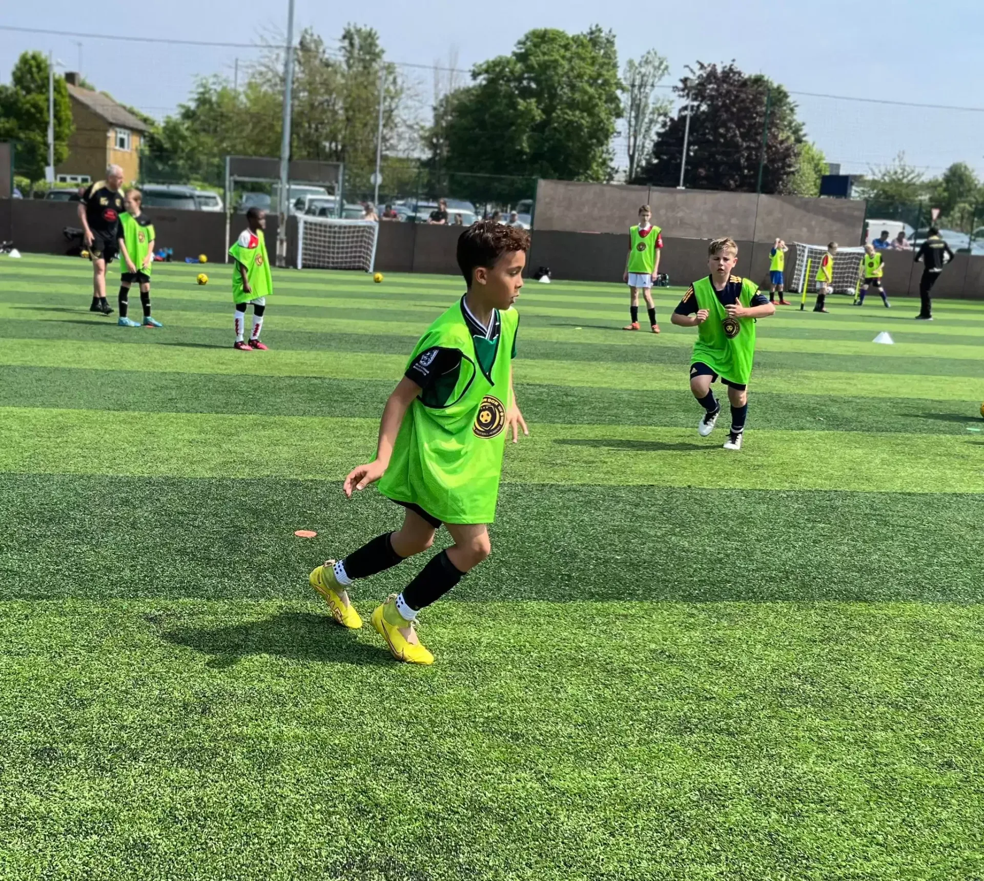 Children playing soccer on a green turf field, with one boy in neon green running in the foreground.