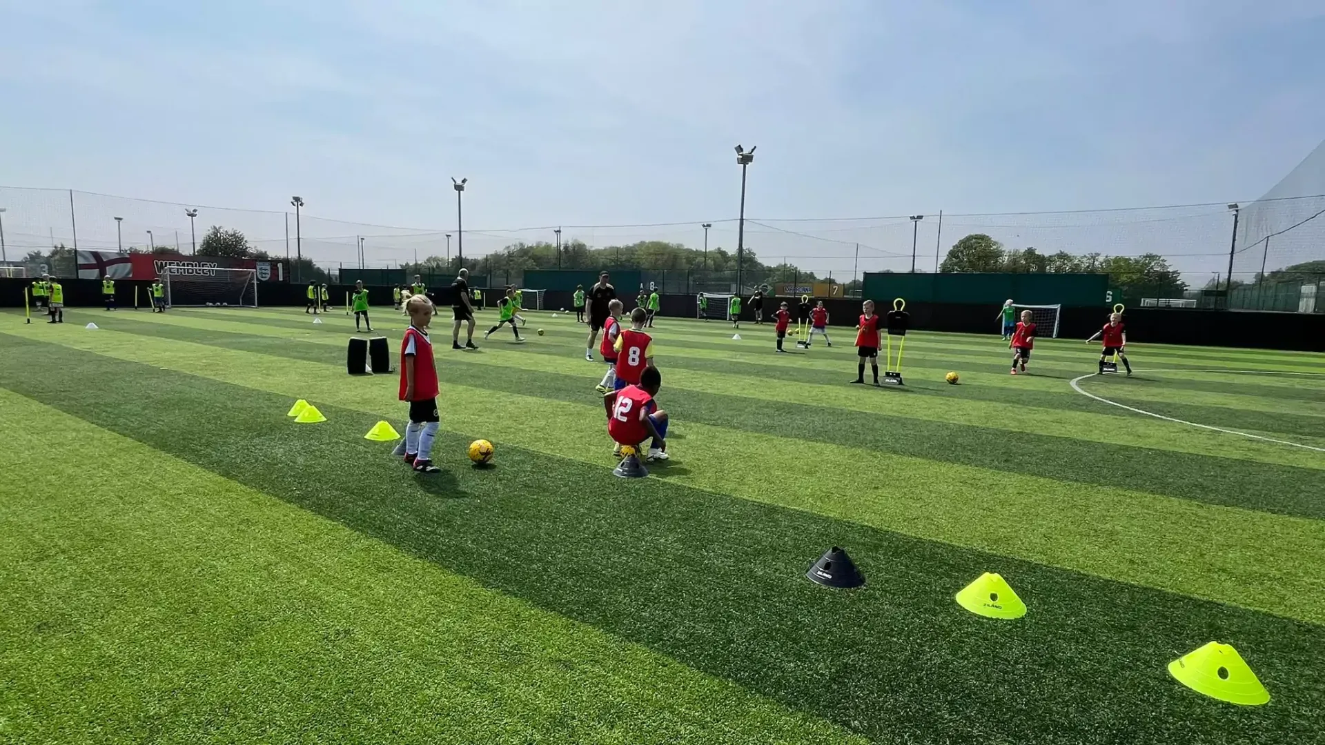 Soccer practice on a green turf field with children in red jerseys and yellow cones lined up.