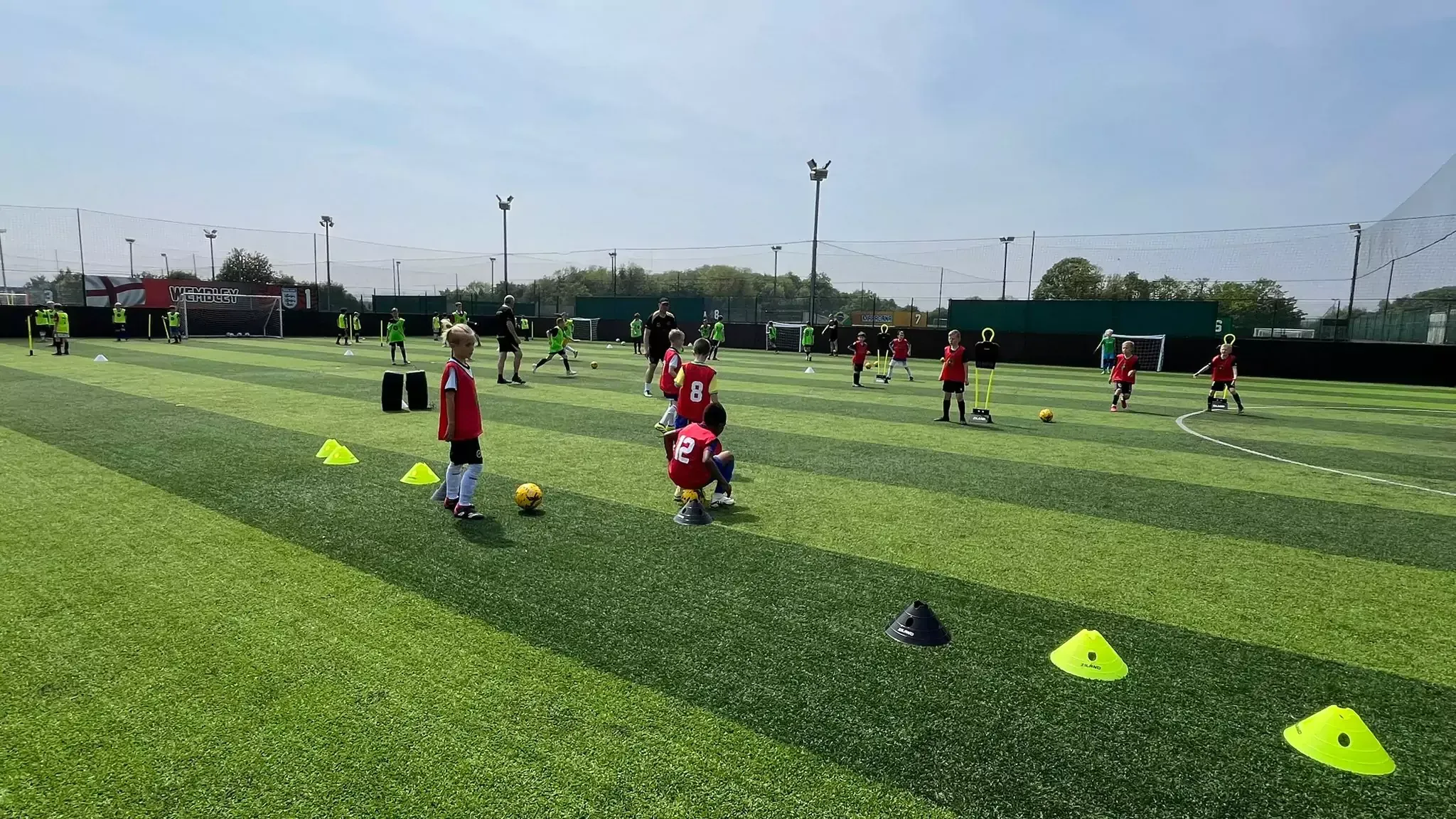 Soccer practice on a green turf field with children in red jerseys and yellow cones lined up.