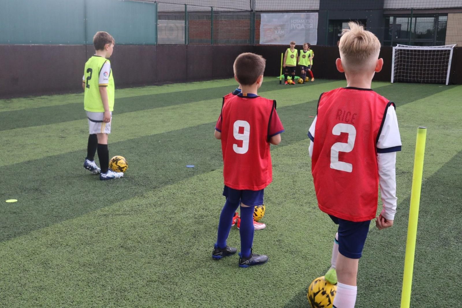 Children in red jerseys wait for a soccer drill on a field, with a coach and goal in the background