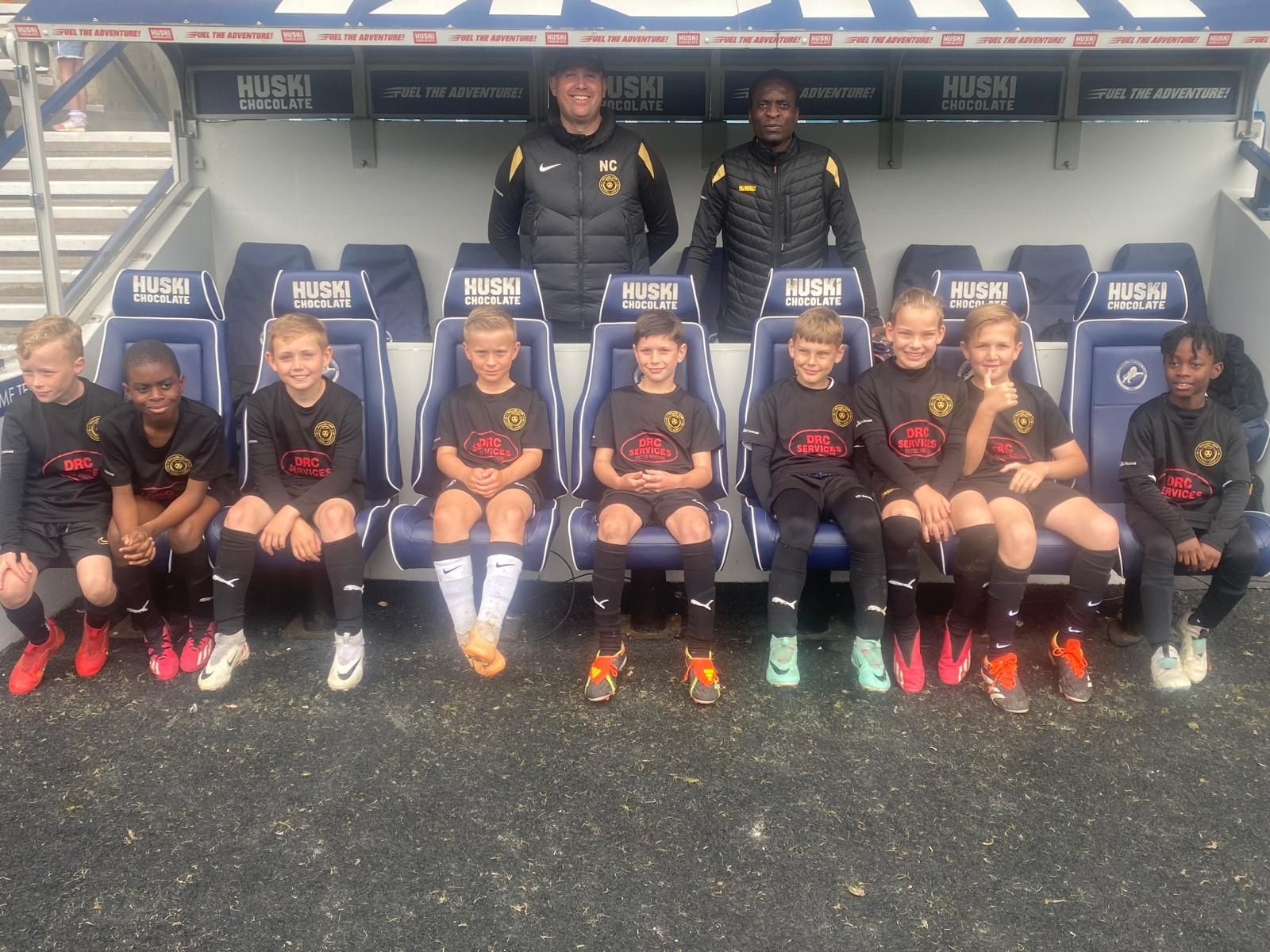 Soccer team posing on bench in a stadium dugout, wearing black uniforms and cleats, with coaches behind them.