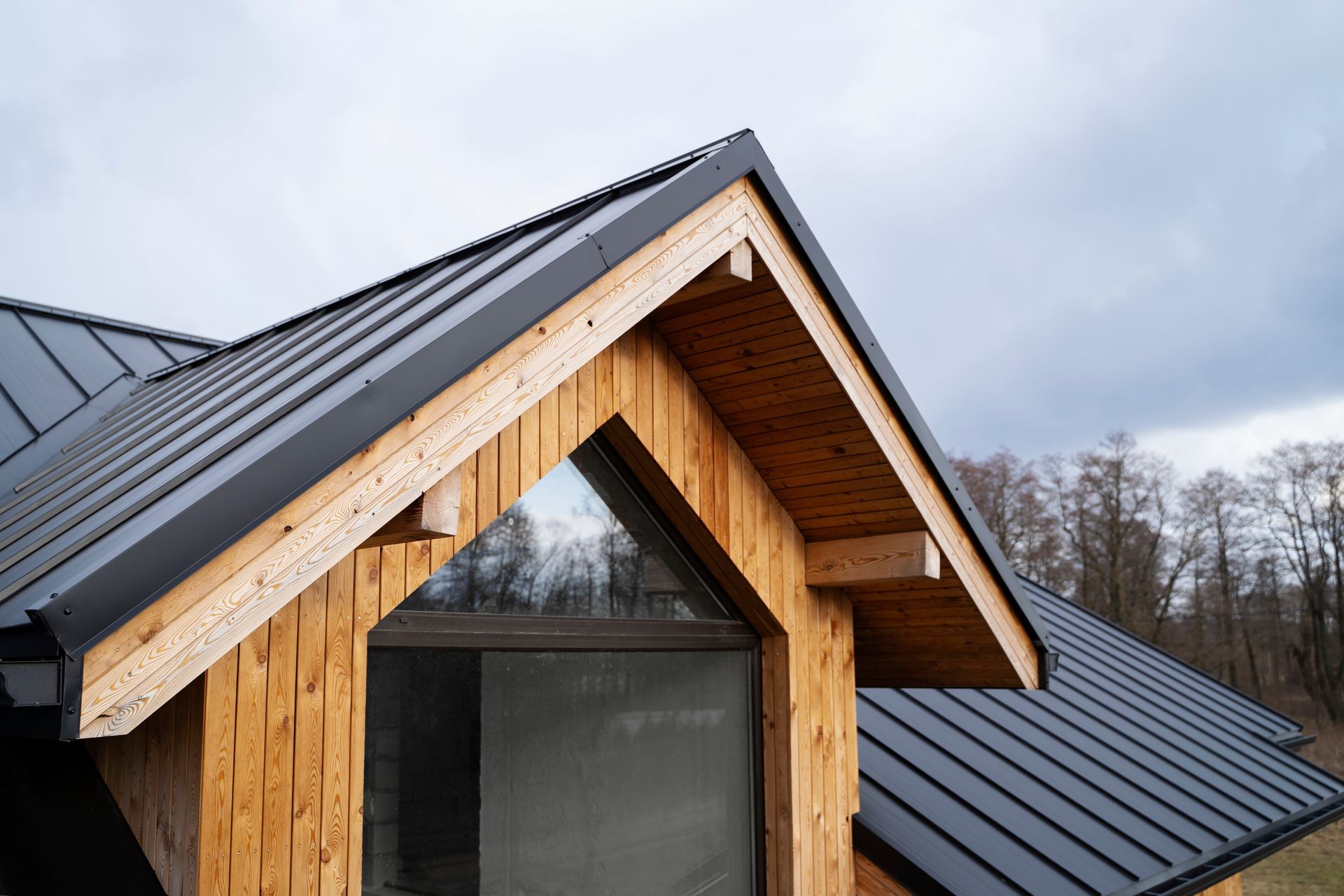 Modern house roof with dark metal panels and a wooden gabled dormer against a cloudy sky