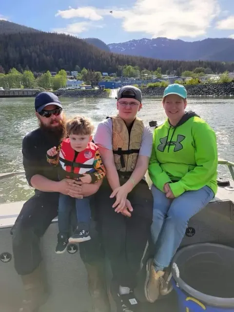 Four people sitting together on a boat, with mountains and a lake in the background.