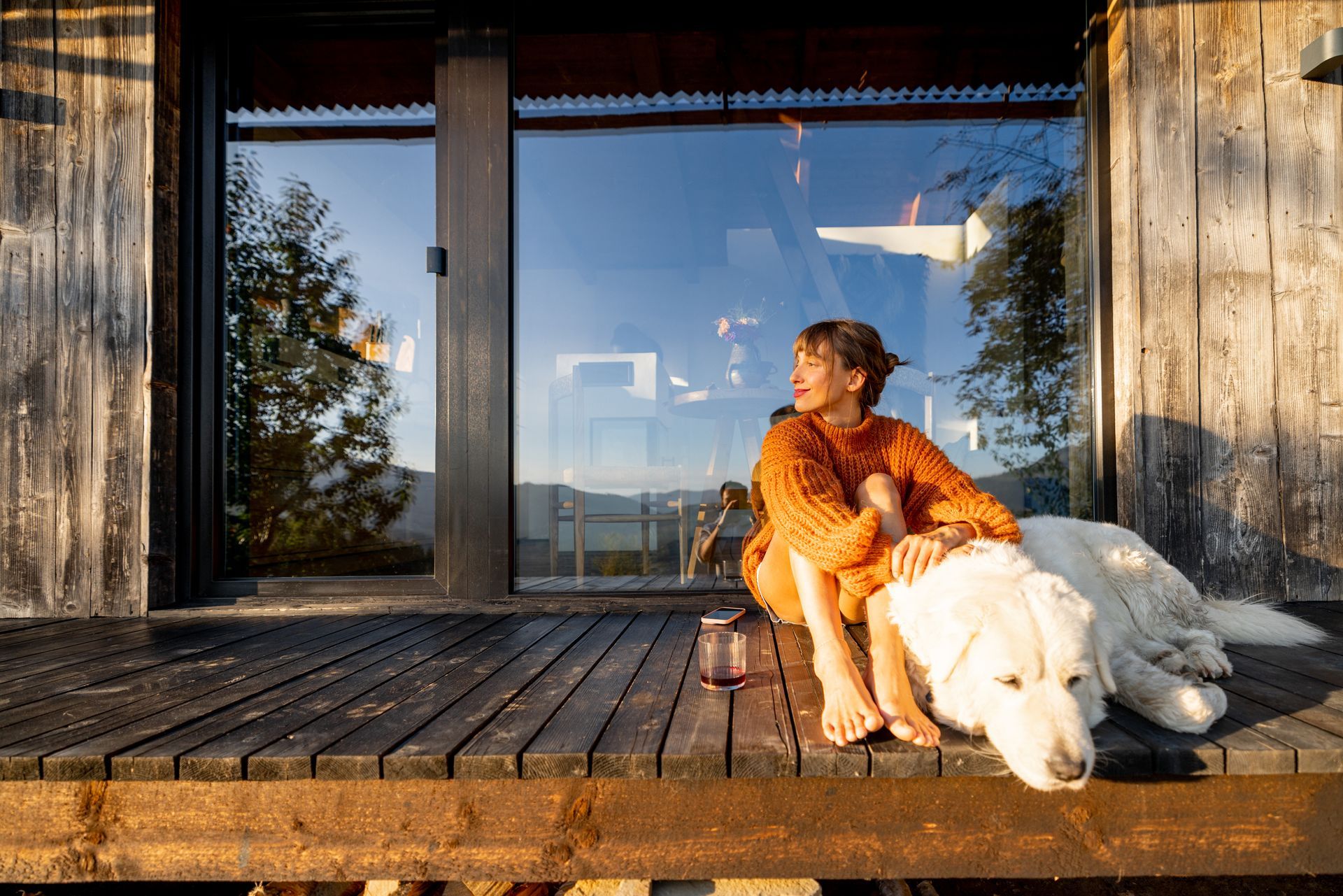 Person in an orange sweater sits on a wooden deck with a white dog beside a glass house.