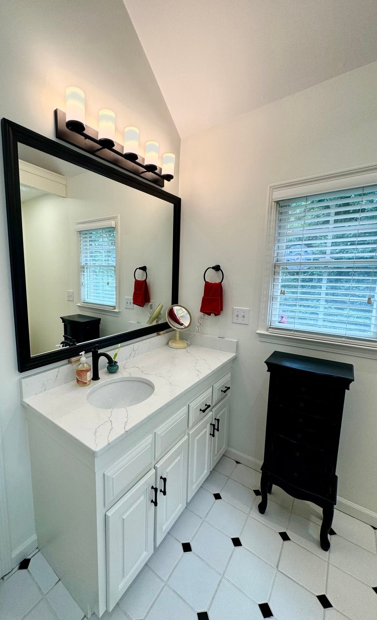 Small white bathroom vanity with mirror, sink, black side cabinet, and window blinds on the right.