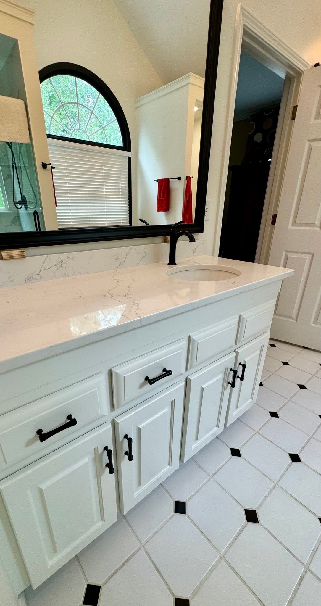 Bathroom vanity with white cabinets, black faucet, round mirror, and tiled floor by an arched window