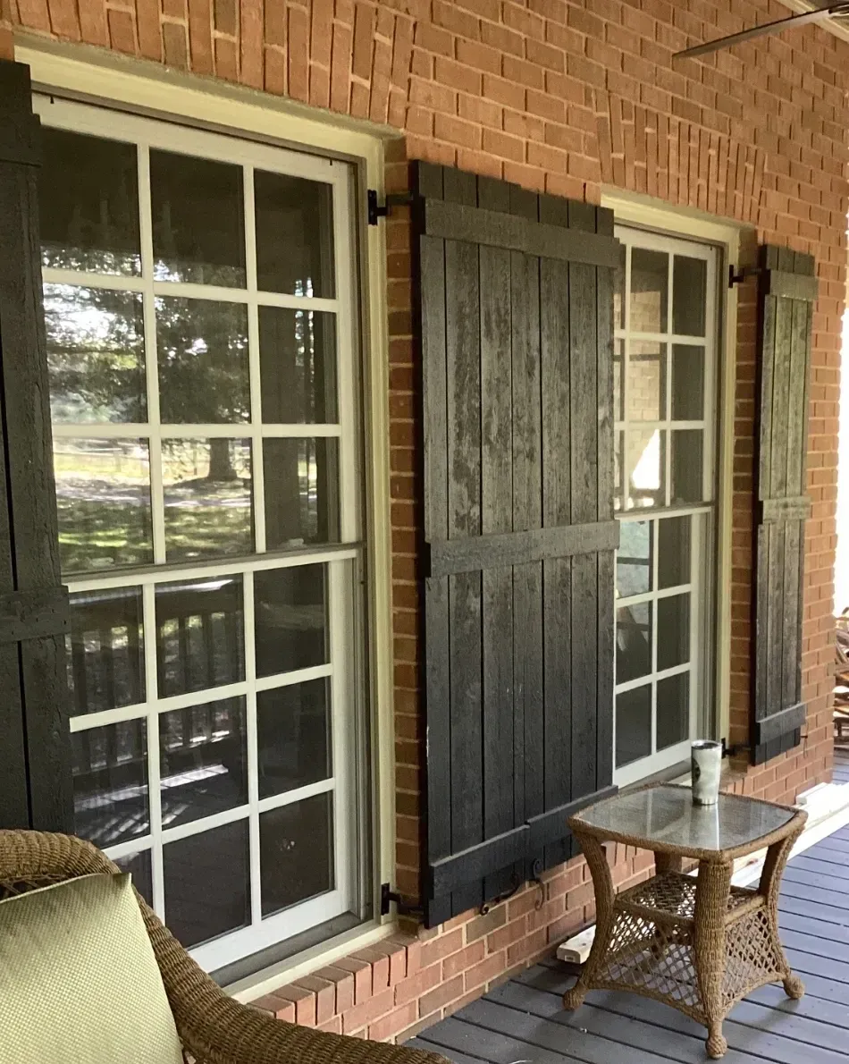 Brick porch with two tall white-trimmed windows, black shutters, and a small wicker table.