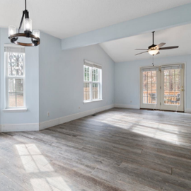 Empty room being painted, with green walls, wood floor, ladder, and a hand painting near a bright doorway. Bright empty living room with gray wood floors, pale blue walls, windows, and a ceiling fan.