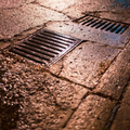 Wet cobblestone sidewalk with two metal storm drains at night under warm streetlight.