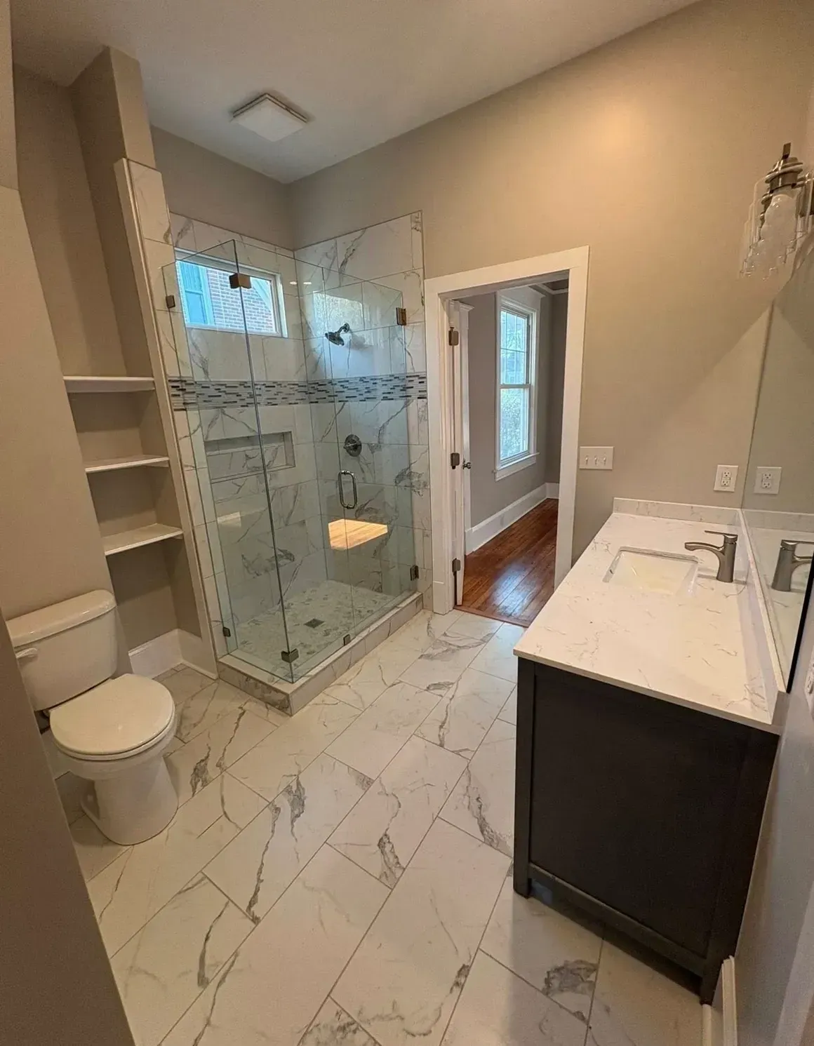 Modern bathroom with glass shower, white marble tile, vanity, toilet, and built-in shelves.