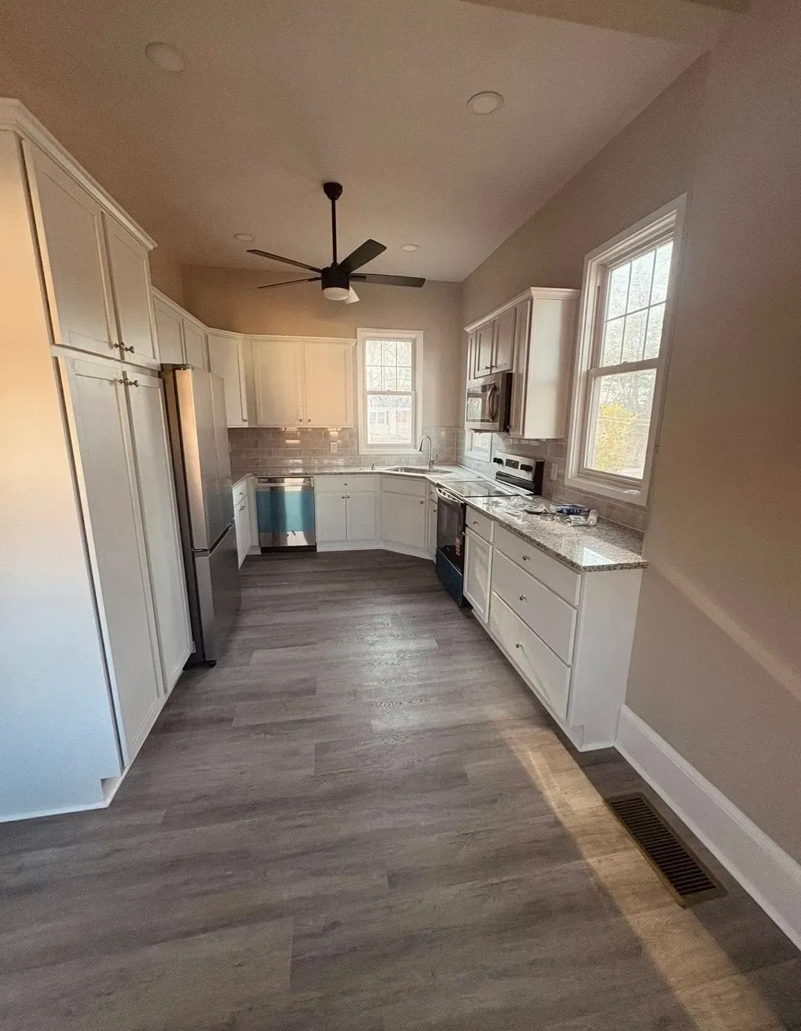 Bright narrow kitchen with white cabinets, gray wood floor, ceiling fan, and windows along one wall