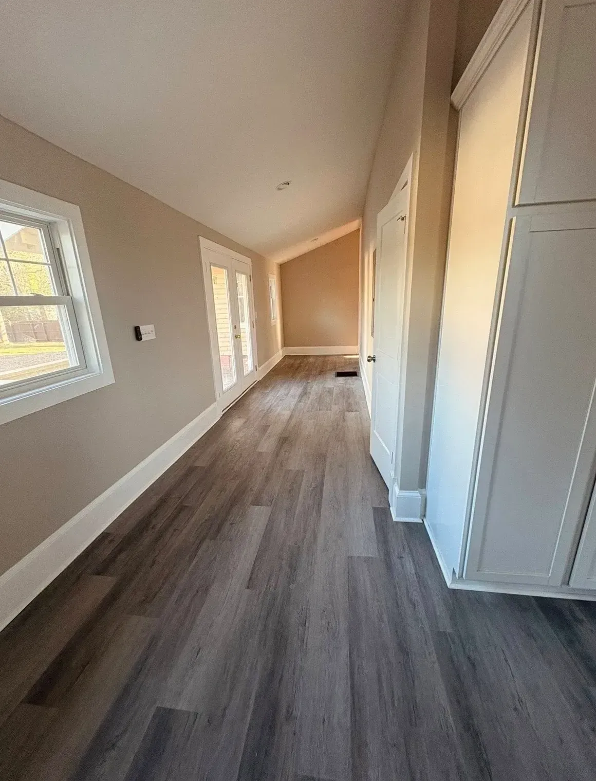 Empty hallway with gray wood floors, beige walls, white trim, and windows along one side