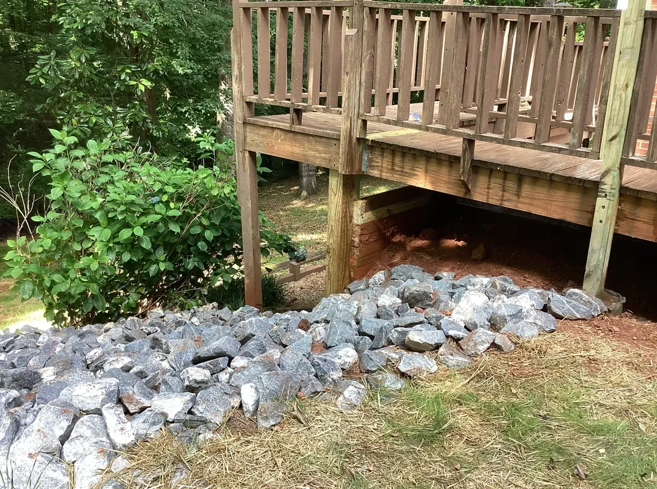 Wooden deck over a rocky drainage bed beside dense green shrubs and trees