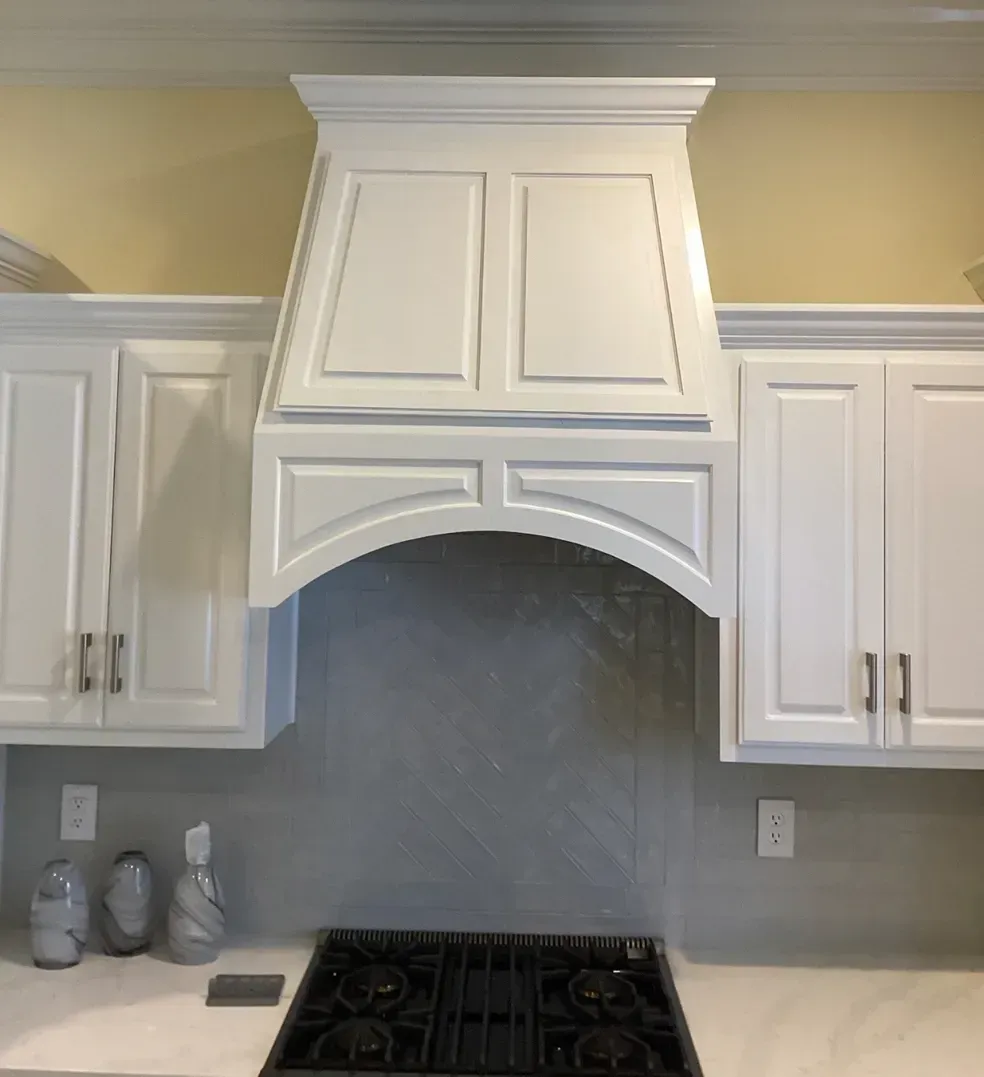 White kitchen range hood with cabinet surrounds above a gas cooktop and gray tile backsplash.