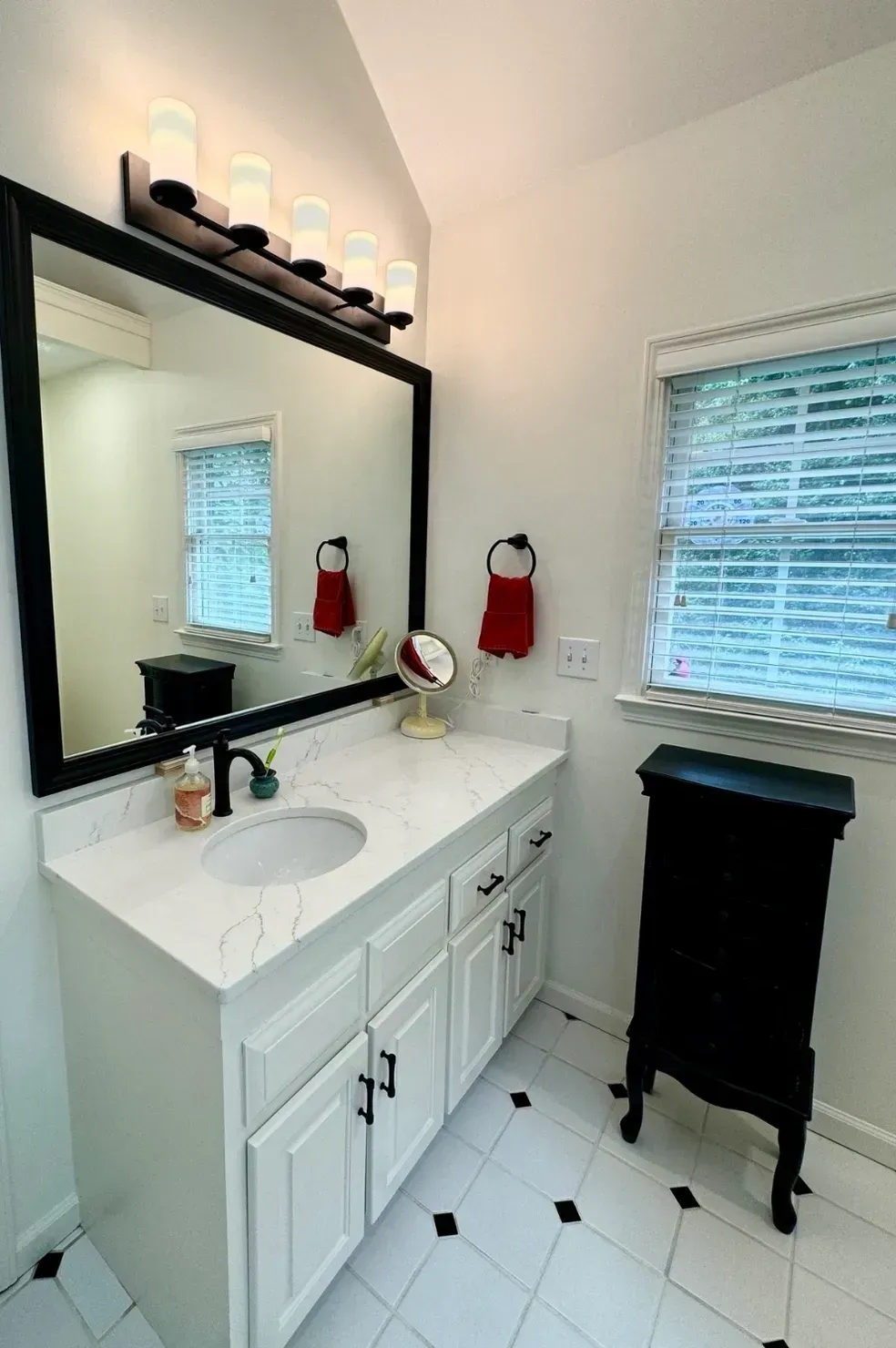White bathroom vanity with double sinks, black-framed mirror, window blinds, and black hamper on tiled floor