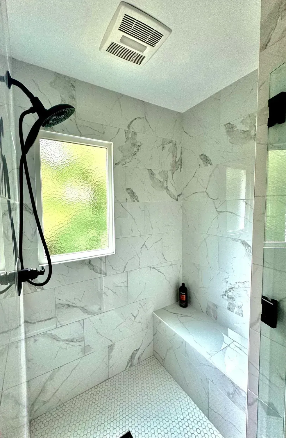 Small marble-tiled shower with a window, rainfall showerhead, and built-in bench.