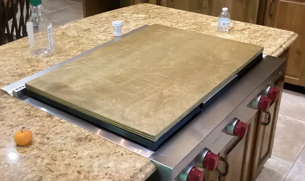 Large square griddle cover on a kitchen stove with granite countertops and red control knobs.