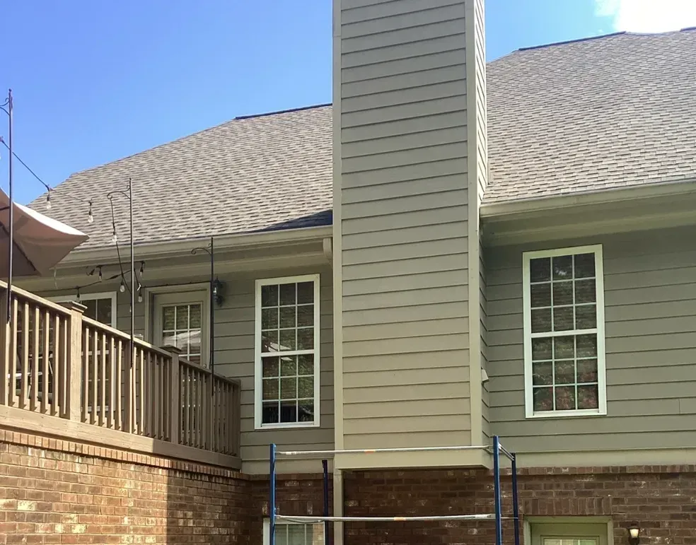 Back of a house with tan siding, brick base, windows, and a central chimney under a shingled roof.