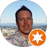 Headshot of a smiling man outdoors with a city skyline in the background and an orange star badge overlay
