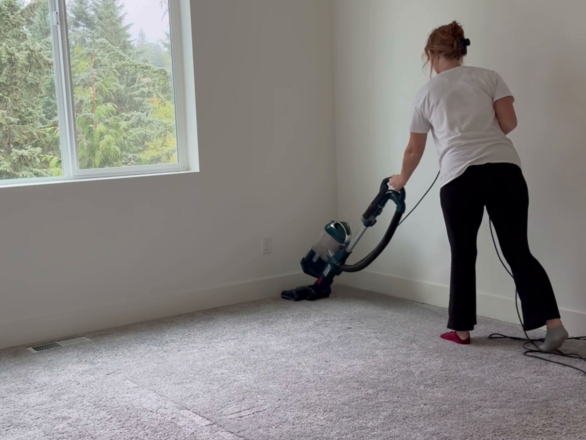 Person vacuuming a carpeted room near a window with a blue vacuum cleaner.