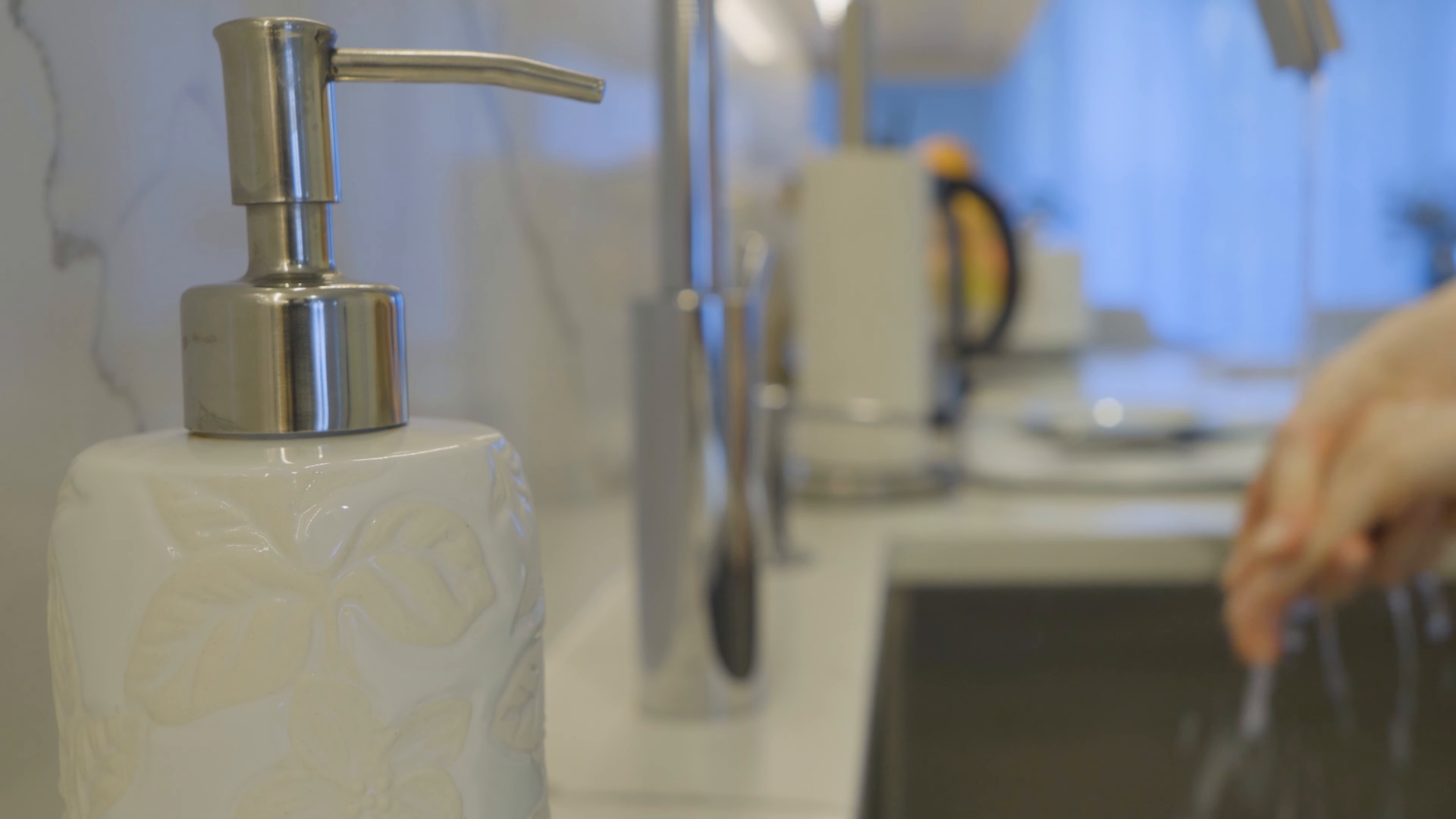 White soap dispenser by a kitchen sink with a kettle and a hand in the background