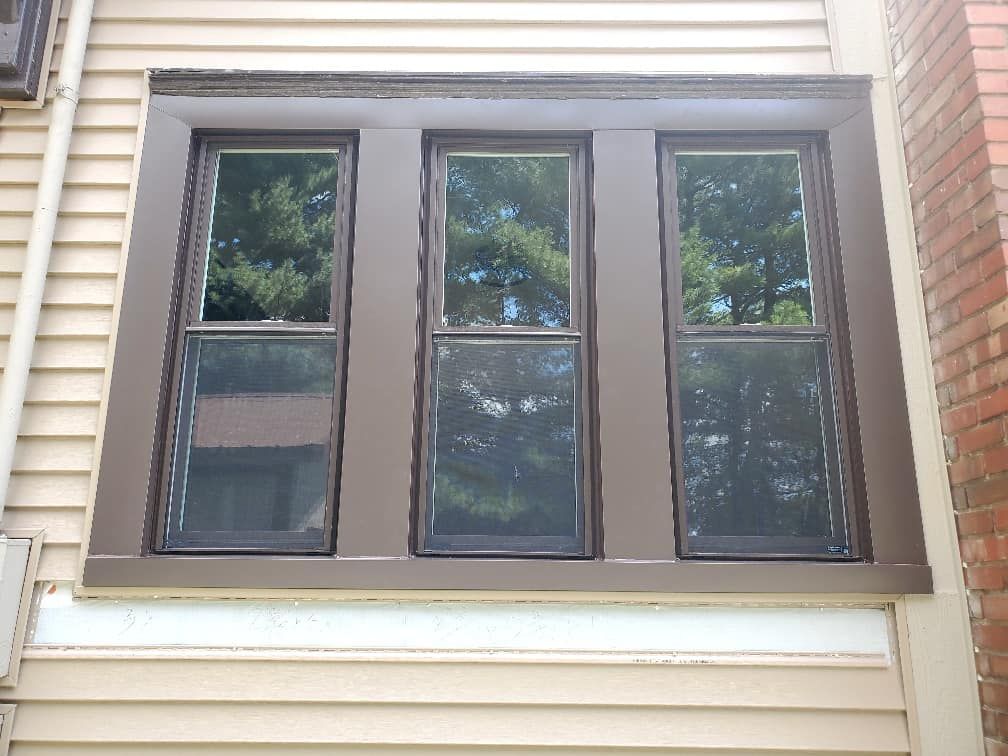 Three brown-framed windows with dark screens and tree reflections on a beige house exterior.