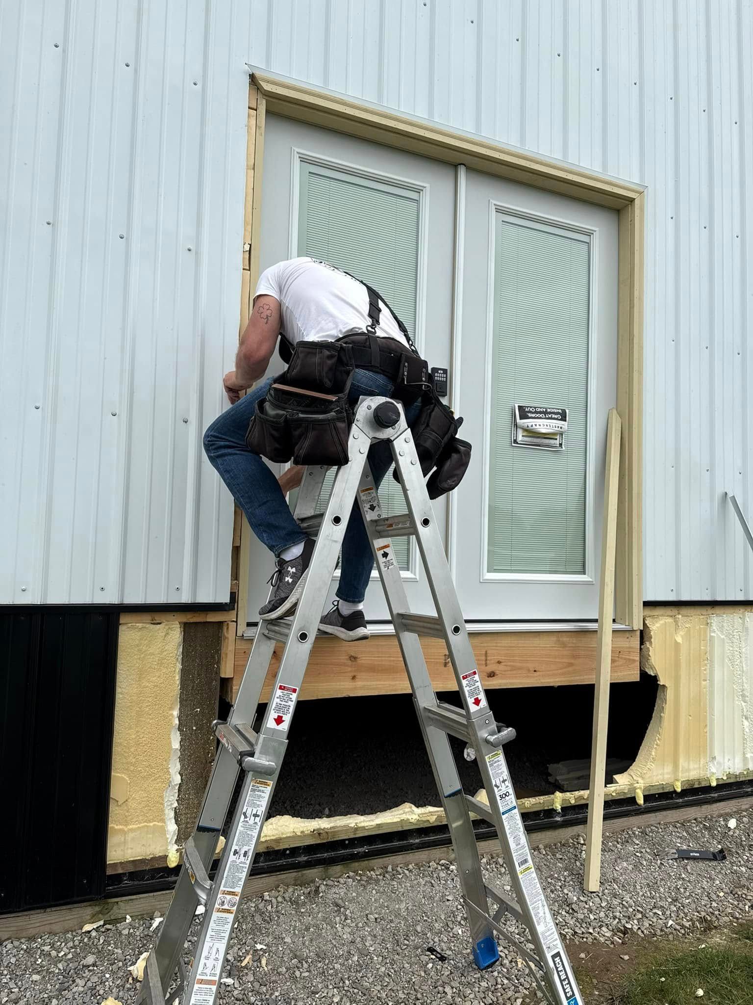 Person installing a door, standing on a ladder. Exterior view of a building with light-colored siding.