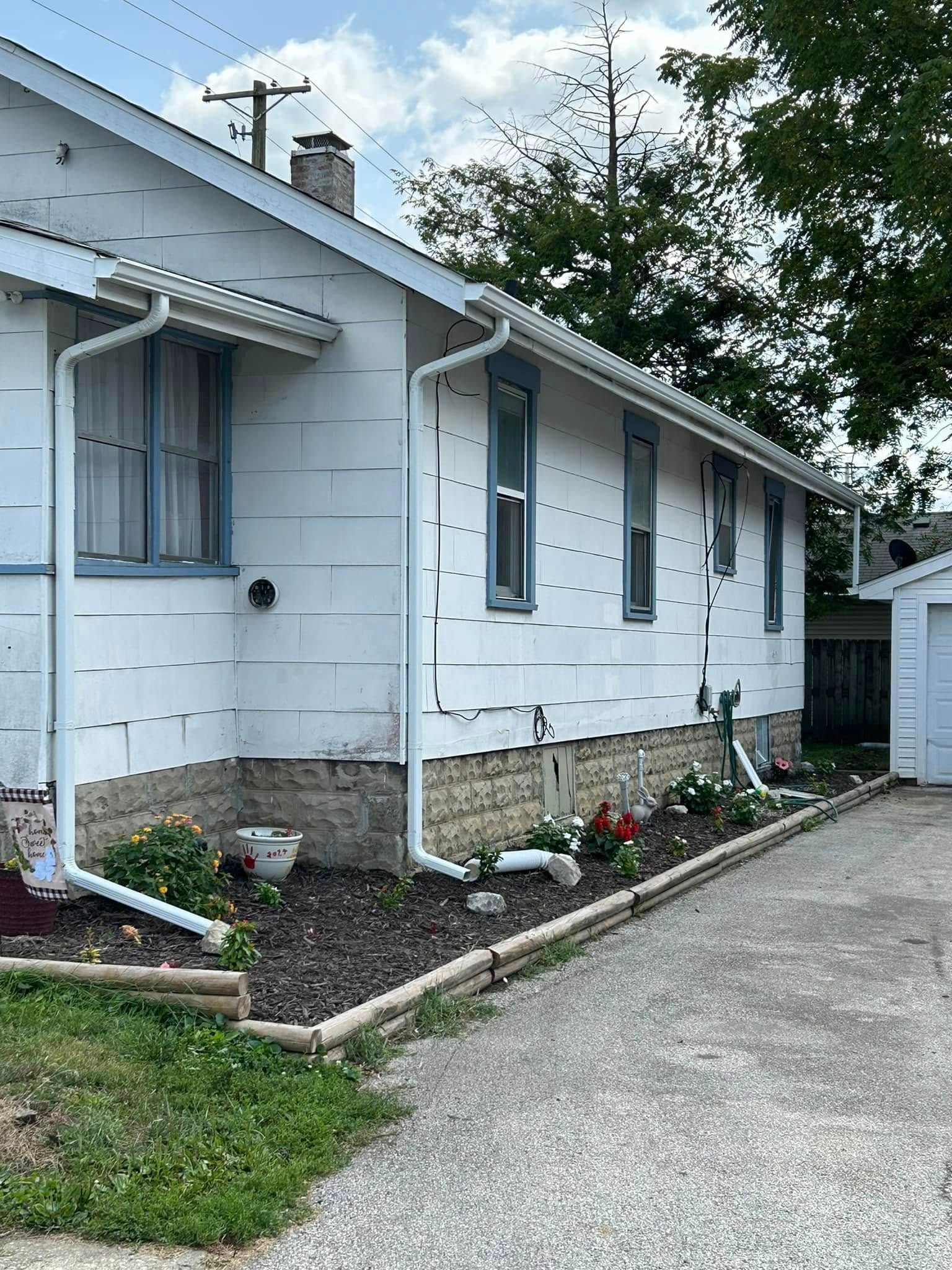 White house with light blue window trim, gutters, and a small flower bed alongside a gravel driveway.