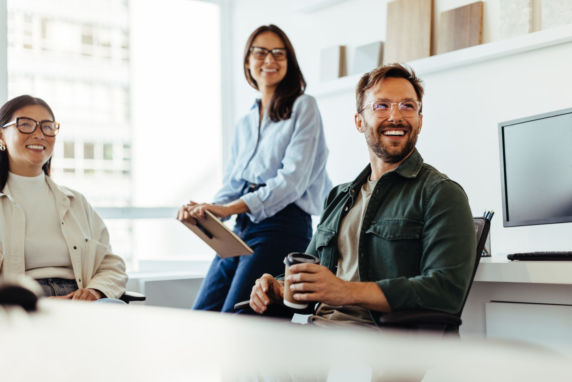 Three coworkers smiling in a bright office, one holding a tablet and another seated at a desk with a monitor