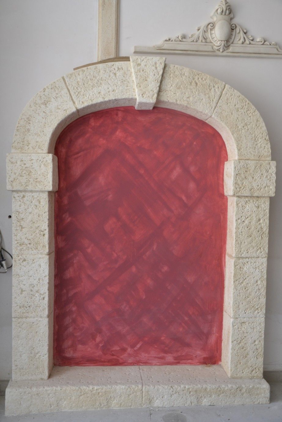 Un arco con una cornice in pietra lavorata che racchiude un inserto rosso opaco e solido, appoggiato a una parete semplice.