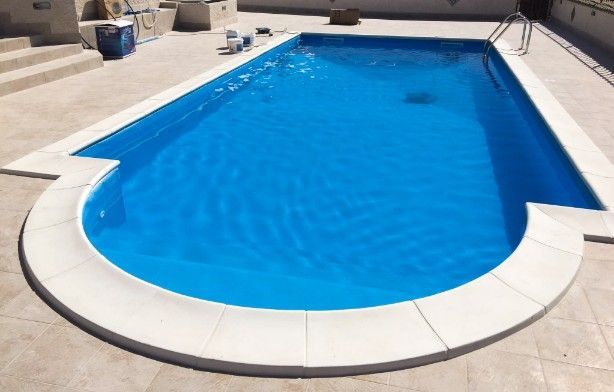 Una piscina rettangolare con estremità arrotondata, circondata da piastrelle in pietra chiara in una giornata di sole.