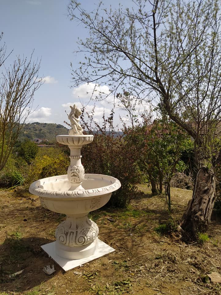 Una fontana in pietra chiara, a più livelli, si trova in un'area esterna con alberi e un terreno collinare sullo sfondo.