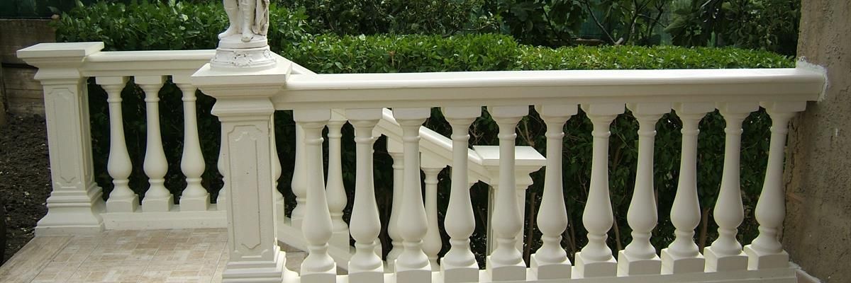 Una balaustra in pietra bianca con fusi decorati incornicia un patio contro una siepe verde.