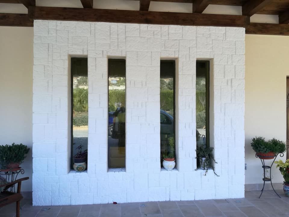 Un muro di pietra bianca con quattro finestre rettangolari verticali, situato sotto un tetto di legno con piante in vaso.