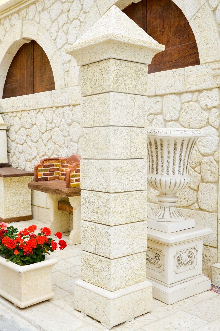 Un pilastro di pietra accanto a un'urna decorativa e a una fioriera con fiori rossi, di fronte a un rustico muro di pietra.