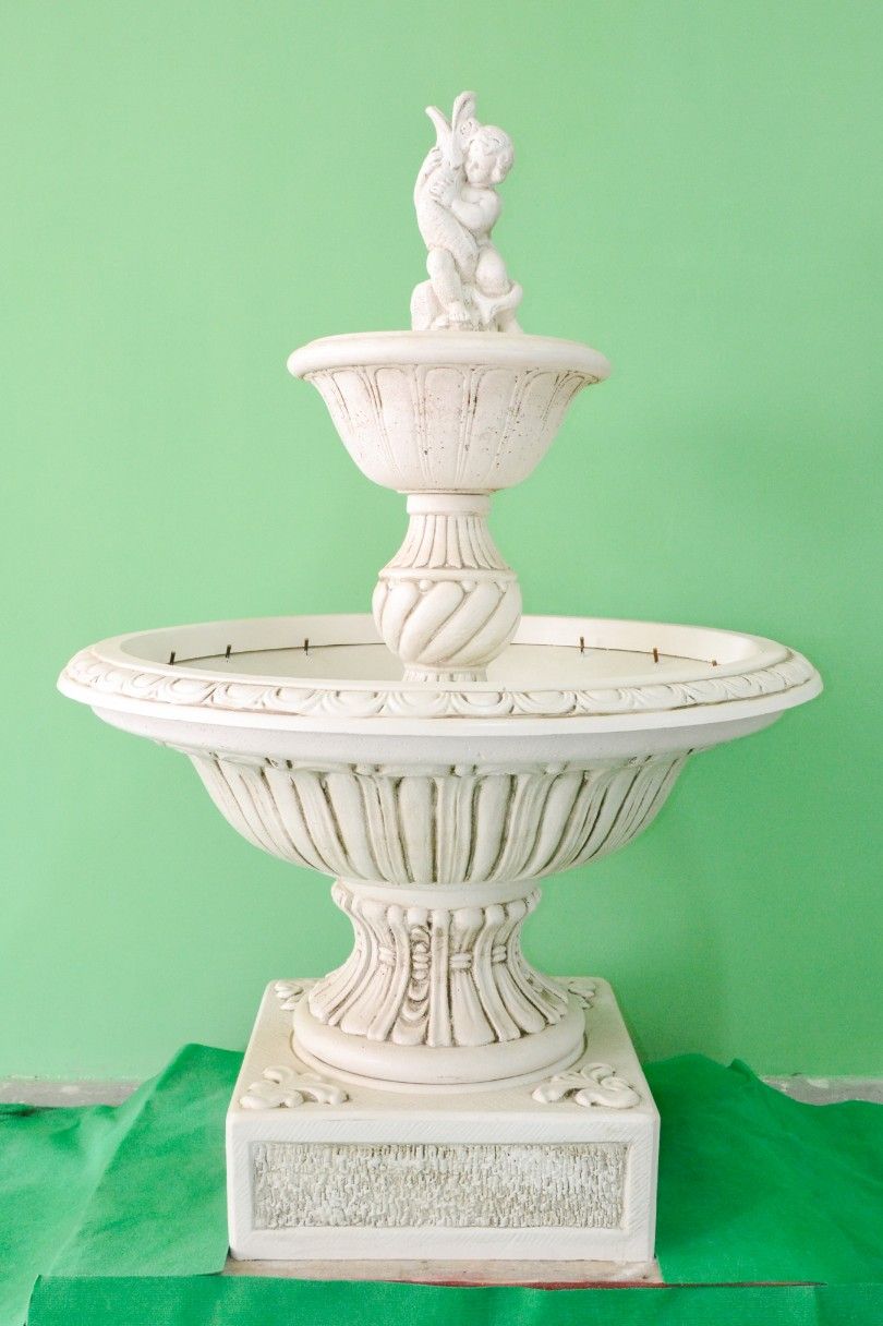 Fontana a più livelli in pietra bianco sporco, con un cherubino decorativo in cima, su uno sfondo verde uniforme.
