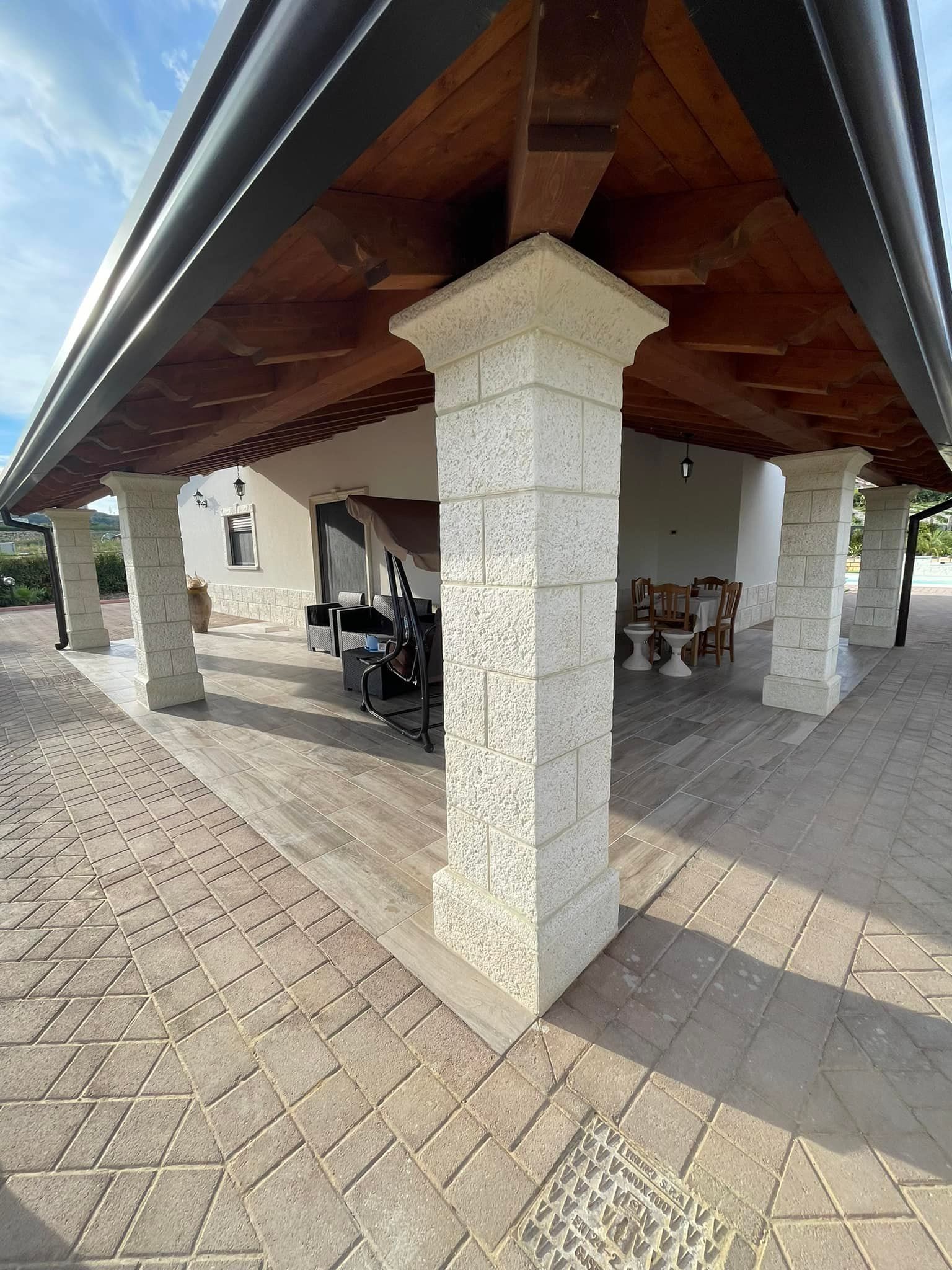 Un patio con pilastri in pietra e tetto in legno si affaccia su un cortile lastricato sotto un cielo luminoso.