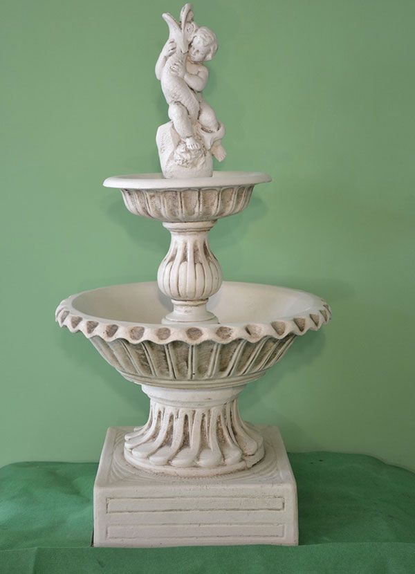 Fontana in pietra a due livelli color bianco sporco, con una figura decorativa di un cherubino, su uno sfondo verde uniforme.