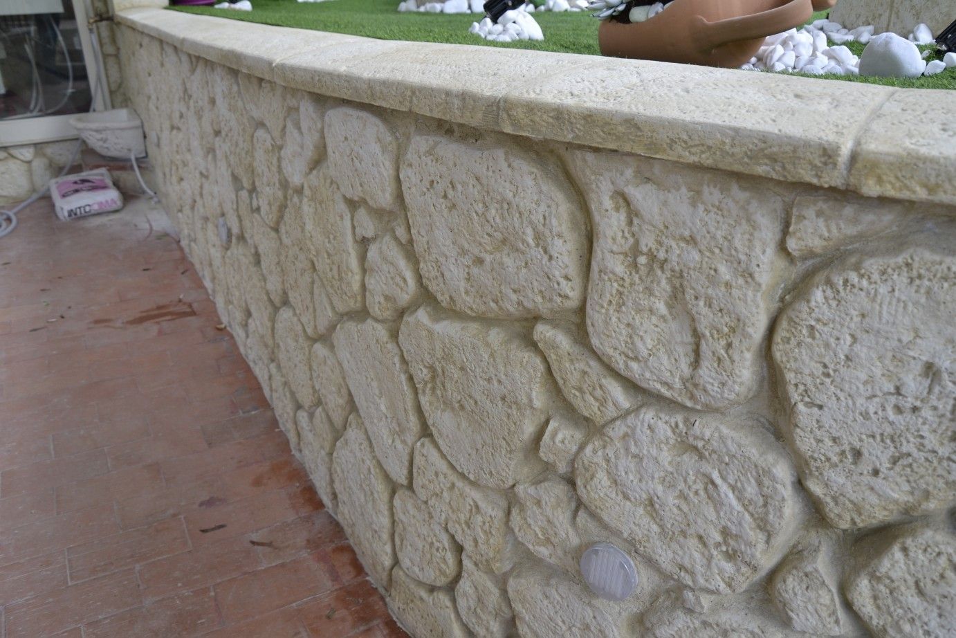 Un muro in pietra grezza beige con un bordo liscio in pietra di copertura, adagiato su un prato e un patio pavimentato.