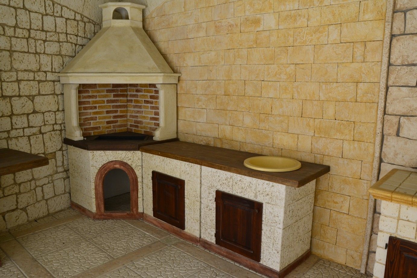 Una cucina esterna in pietra con griglia angolare, camino, piano di lavoro, lavello e ante in legno.