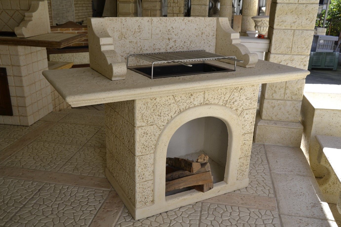Barbecue da esterno in pietra beige con un arco centrale per riporre la legna e una griglia metallica sul piano di lavoro.