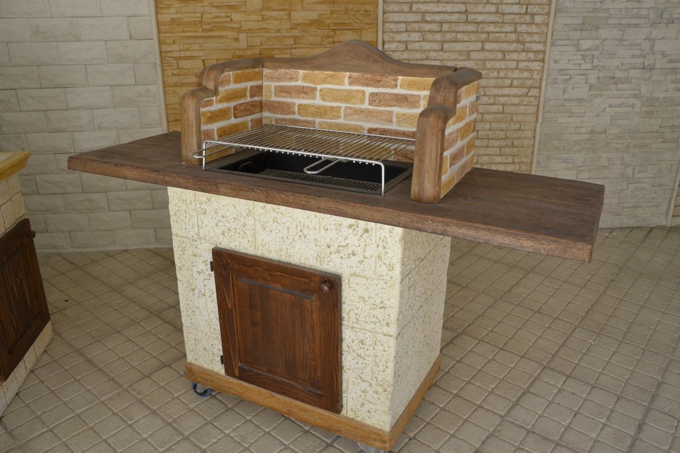 Barbecue mobile in muratura per esterni con paraschizzi in mattoni, porta in legno scuro e base con texture in pietra.
