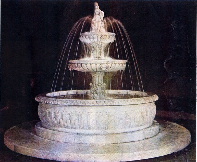 Una fontana in pietra bianca a tre livelli con acqua che scorre, posta su uno sfondo scuro.