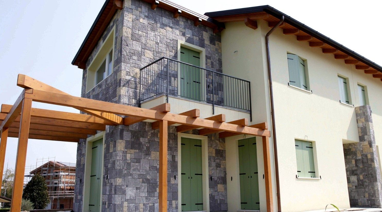 Edificio a due piani con rivestimento in pietra, pareti chiare, persiane verdi e un pergolato in legno che copre l'ingresso.